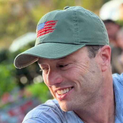 Man wearing a green cap with an American flag design, sitting outdoors.
